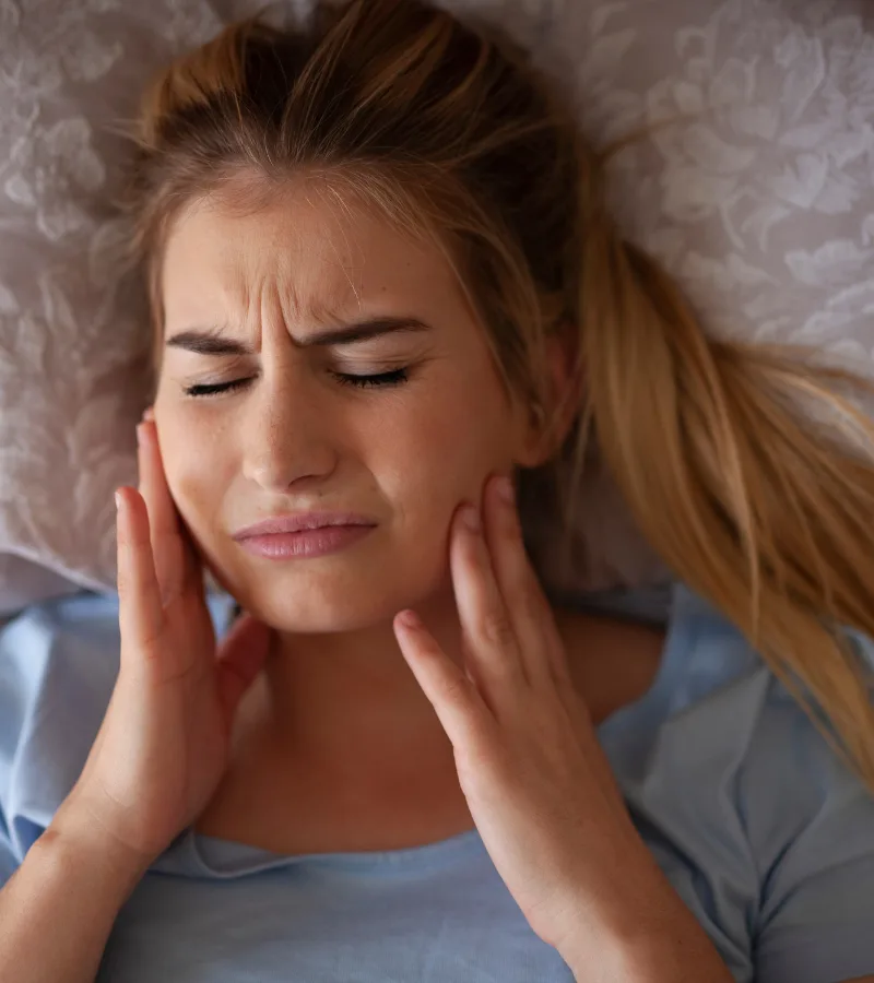 Young woman in pain holding her cheeks, showing common symptoms of teeth grinding (bruxism) – treated at Aesthetique Dental Care in Leeds.